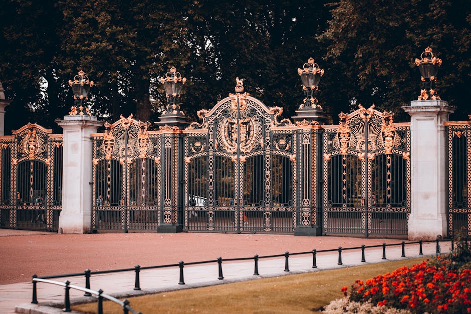 An ornate wrought iron gate with intricate gold detailing and decorative scrollwork, set between tall white stone pillars topped with elaborate gold finials. Behind the gate, mature trees with dense f