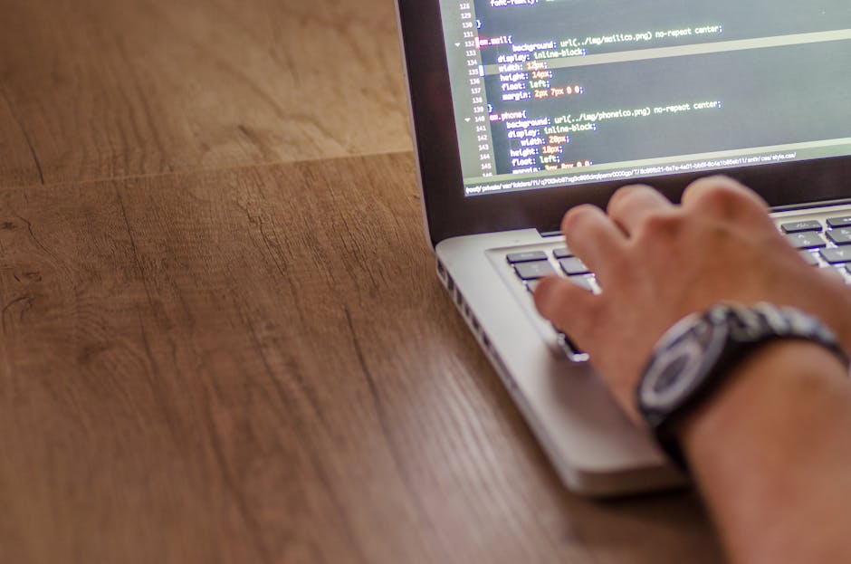 The image shows a person's hand typing on a laptop placed on a wooden desk. The laptop screen displays lines of code with various colors highlighting syntax, suggesting a programming or web developmen