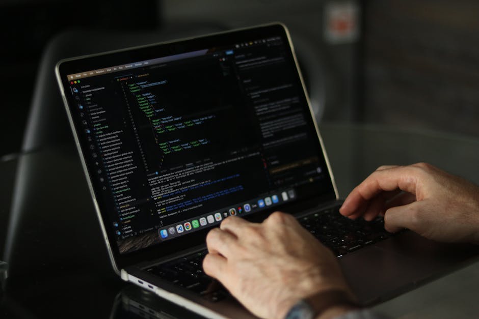 The image shows a person's hand typing on a laptop placed on a wooden desk. The laptop screen displays lines of code with various colors highlighting syntax, suggesting a programming or web developmen